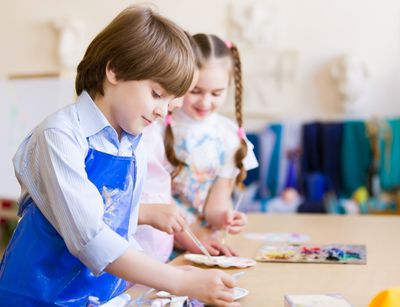 Im Vordergrund ist auf diesem Foto ein circa 10 jähriger Junge im Kunstunterricht abgebildet. Er trägt eine blaue Schürze und malt im stehen am Tisch ein Bild. Im Hintergrund steht ein Mädchen mit geflochtenen Zöpfen.