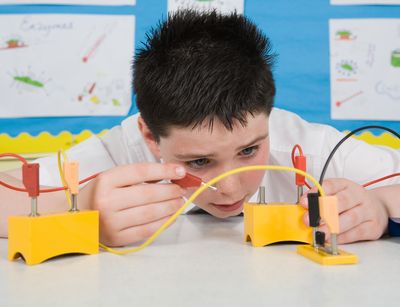 Auf dem Foto ist ein circa 11 jähriger Junge im Physikunterricht abgebildet. Er hat einen selbst gebauten Schaltkreis vor sich auf dem Tisch stehen und probiert sehr konzentriert die elektrische Ladung aus.