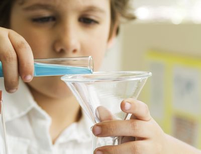 Auf dem Foto wird ein circa 11 Jahre alter Junge abgebildet, der im Chemieunterricht eine blaue Flüssigkeit in ein Reagenzglas füllt.