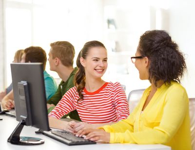 Auf dem Foto sind im Vordergrund zwei ca 15 jährige Schülerinnen im Informatikunterricht abgebildet, die zueinander gewandt miteinander lachen. Vor ihnen steht ein Computerbildschirm und im Hintergrund sind drei weitere Schüler an einem Computer.