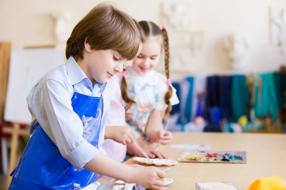 zur Vergrößerungsansicht des Bildes: Im Vordergrund ist auf diesem Foto ein circa 10 jähriger Junge im Kunstunterricht abgebildet. Er trägt eine blaue Schürze und malt im stehen am Tisch ein Bild. Im Hintergrund steht ein Mädchen mit geflochtenen Zöpfen.