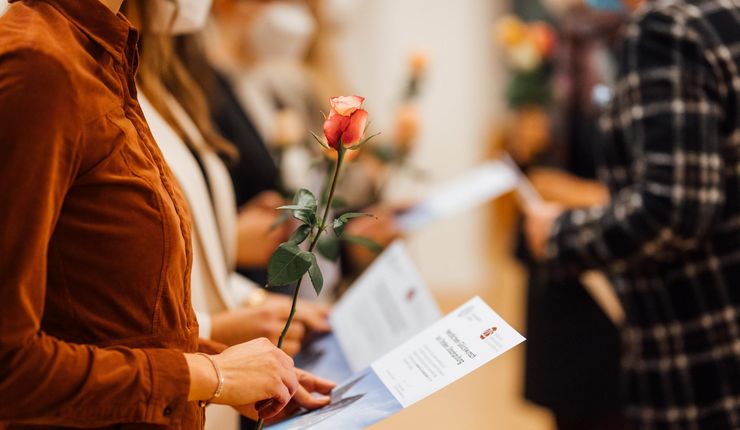 Die linke Seite des Fotos zeigt teilweise unscharf zu erkennende Menschen ohne Gesichter, die jeweils eine Rose und eine Urkunde in den Händen halten. Nur unscharf zu erkennen befindet sich an der rechten Seite des Bildes eine Person (ohne Gesicht) im karierten Jackett.