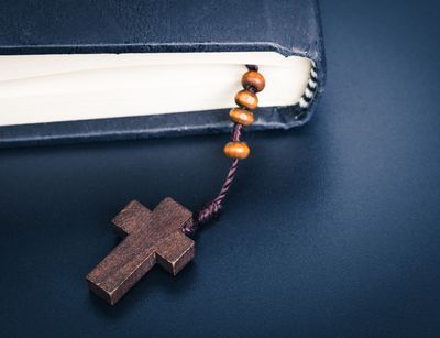 Auf dem Foto ist der untere Abschnitt einer zugeklappten Bibel abgebildet, aus dem eine Kette mit einem Holzkreuz und Perlen herausguckt.