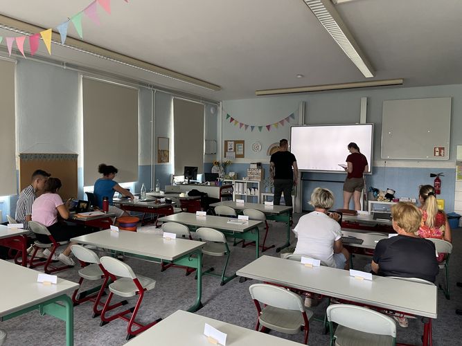Das Foto zeigt ein Klassenzimmer aus einer hinteren Perspektive Richtung Smartboard. Vereinzelt sitzen Erwachsene an den Schüler:innentischen. Zwei Personen stehen vorn an der digitalen Tafel. 