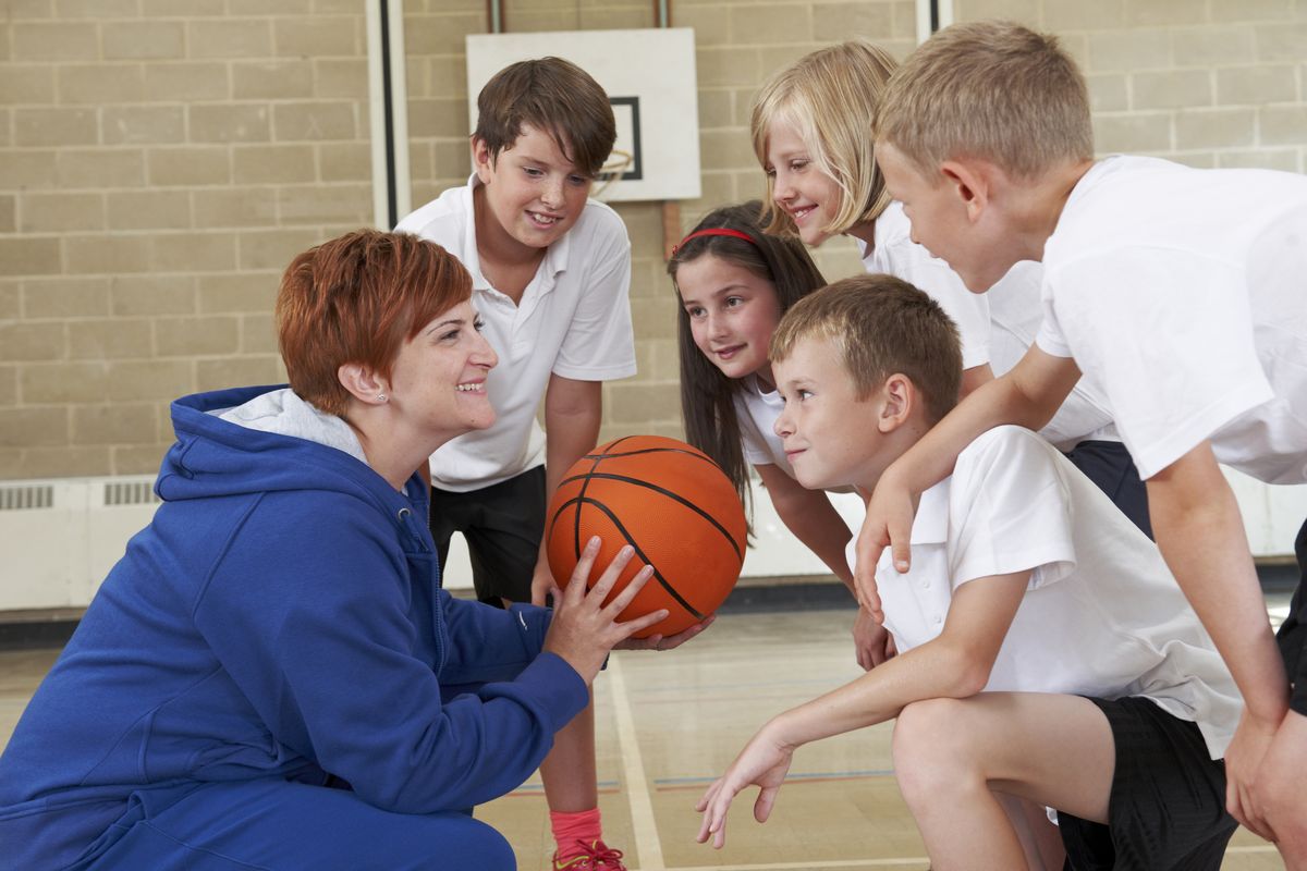 zur Vergrößerungsansicht des Bildes: Auf dem Foto ist links eine Lehrerin mit kurzen Haaren im Sportunterricht abgebildet. Sie hockt vor fünf Kindern und hält einen Basketball in der Hand und erklärt lachend etwas.