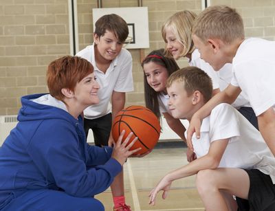 Auf dem Foto ist links eine Lehrerin mit kurzen Haaren im Sportunterricht abgebildet. Sie hockt vor fünf Kindern und hält einen Basketball in der Hand und erklärt lachend etwas.