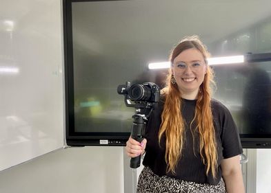 Laura Kirste, Lehramtsstudentin und wissenschaftliche Hilfskraft am Zentrum für Lehrer:innenbildung und Schulforschung (ZLS). Foto: Rosa Sommer Zu sehen ist Laura Kirste, in der Hand hält sie ein Stativ mit einer Kamera.