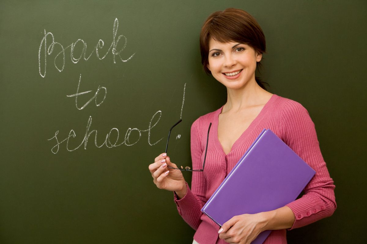 Junge Frau mittleren Alters steht vor einer Tafel und lächelt in die Kamera. An der Tafel steht in weißer Kreideschrift Back to School geschrieben.