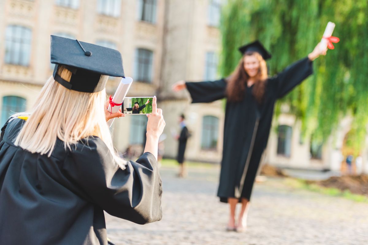 zur Vergrößerungsansicht des Bildes: Zwei Universitätsabsolventinnen in typischer Abschlussfeierkleidung, mit Diplom in der Hand, die eine fotografiert die andere mit ihrem Handy. Foto: Colourbox 