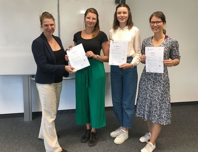 Auf dem Bild sind drei Studentinnen und eine Dozentin zu sehen, die ein Zertifikat in der Hand halten. Sie stehen in einem Seminarraum vor einer Tafel. 