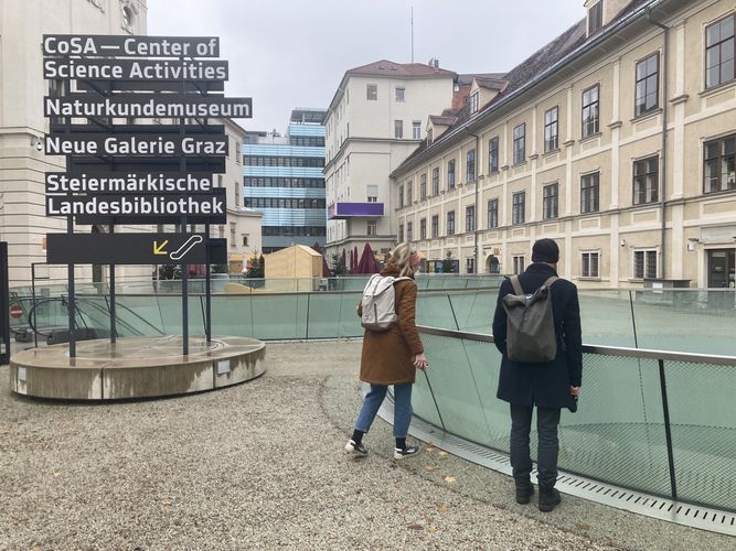 Das Bild zeigt eine Anzeigetafel mit der Auflistung Grazer Museen. Zwei Menschen stehen mit dem Rücken zum Betrachter im Bild.