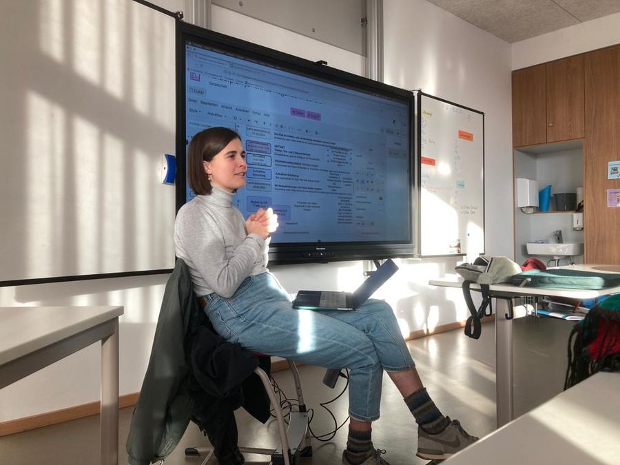 zur Vergrößerungsansicht des Bildes: Sarah Bischof sitzt mit Laptop auf dem Schoß vor einer digitalen Tafel und hält einen Vortrag. 