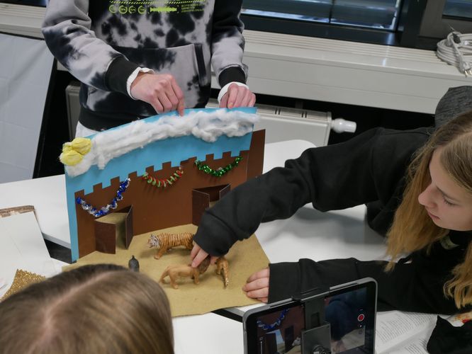 Eine Schülerin arrangiert Löwen- und Tiegerfiguren für eine Szene aus "Der Handschug" von Freidrich Schiller. Ein weiterer Schüler hält die Kulisse fest. Im Vordergrund steht ein Tablet, das die Szene aufnimmt. 