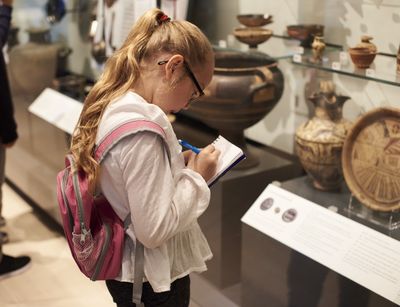 Auf dem Foto ist ein circa 10 jähriges Mädchen mit langen Haaren und einem Rucksack abgebildet, welches sich im Museum alte Vasen und Teller anschaut und sich Notizen macht.