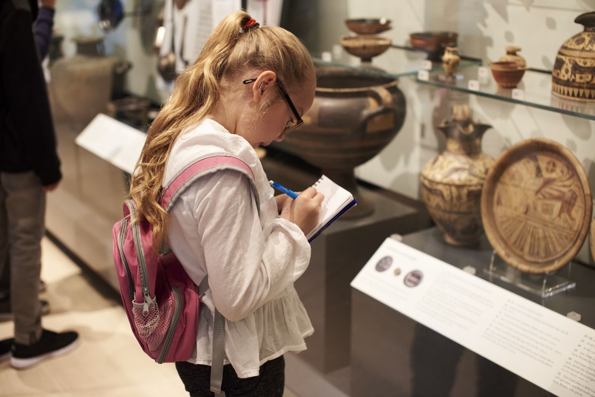 zur Vergrößerungsansicht des Bildes: Auf dem Foto ist ein circa 10 jähriges Mädchen mit langen Haaren und einem Rucksack abgebildet, welches sich im Museum alte Vasen und Teller anschaut und sich Notizen macht.