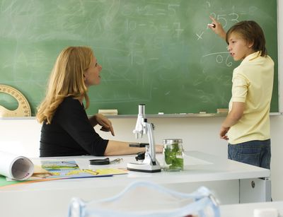 Auf dem Foto ist rechts ein circa 12 jähriger Junge an der Tafel abgebildet. Er schreibt eine Rechenaufgabe an die Tafel und schaut fragend zur Lehrerin. Die Lehrerin sitzt links im Bild am Tisch und schaut zum Jungen.