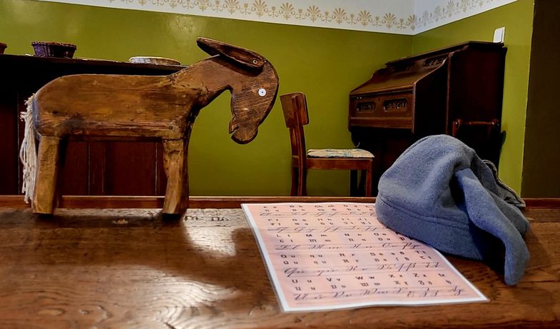 Zu sehen ist ein Holzesel, einem Kinderspielzeug ähnelnd, sowei ein mit Holz verkleidetes Klavier im Hintergrund vor einer grün gefärbten Wand.