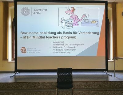Auf dem Bild ist eine weiße Projektionswand zusehen. Auf dieser Ist das Titelbilld der Präderntaiotn für den Kurs Mindful Teacher Program abgebildet. Davor steht ein schwarzer Stuhl. 