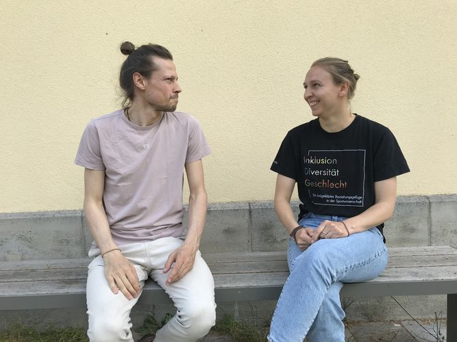 Ein junger Mann auf der linken Bildseite und eine junge Frau auf der rechten Bildseite sitzen sich zugewandt auf einer Bank vor einem Gebäude und sind in einem Gespräch vertieft.