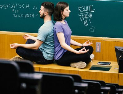 Farbfoto: Eine Frau und ein Mann meditieren in einem Hörsaal vor einer Tafel. Sie sitzen dabei im Schneidersitz auf dem Pult des Dozierenden. Sie haben die Augen geschlossen und sitzen Rücken an Rücken.uf einem Tisch