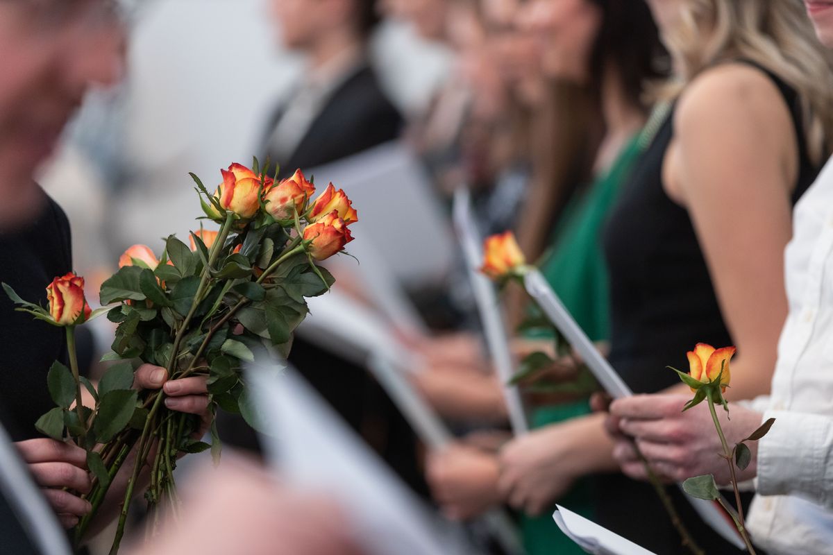 zur Vergrößerungsansicht des Bildes: Das Foto zeigt eine Reihe vom Menschen, die nebeneinander stehen und deren Gesichter unkenntlich sind. Sie halten Mappen und Rosen in den Händen. Weitere nicht erkennbare Personen stehen ihnen gegenüber, die eine hält mehr als fünf Rosen in der Hand. 