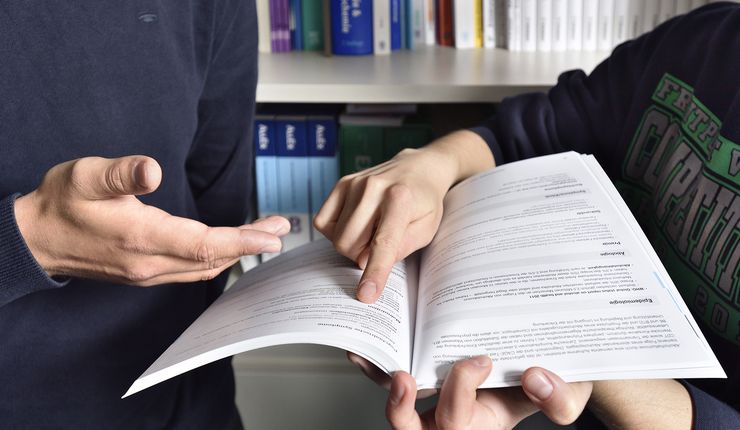 Zu sehen sind die Hände zwei stehender Personen, die in einem Buch nachlesen