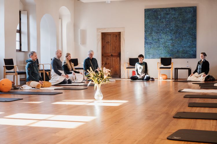 Es ist der Probsteisaal des Klosterhof St. Afra zu sehen. Es sitzen Menschen auf schearen Matten oder orangenen Meditationskissen. Die Menschen sitzen im Kreis. 