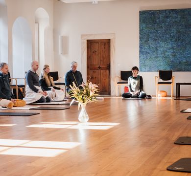 Es ist der Probsteisaal des Klosterhof St. Afra zu sehen. Es sitzen Menschen auf schearen Matten oder orangenen Meditationskissen. Die Menschen sitzen im Kreis. 