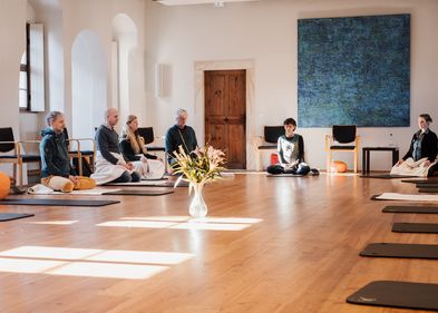 Es ist der Probsteisaal des Klosterhof St. Afra zu sehen. Es sitzen Menschen auf schearen Matten oder orangenen Meditationskissen. Die Menschen sitzen im Kreis. 