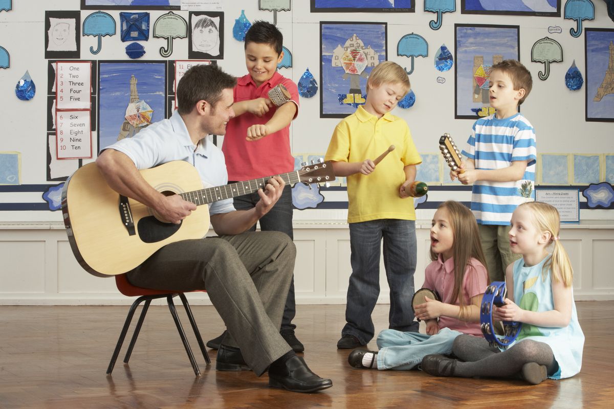 zur Vergrößerungsansicht des Bildes: Links auf dem Bild ist ein junger Lehrer, der Gitarre spielt abgebildet. Neben ihm stehen drei Jungs und sitzen zwei Mädchen, die circa 8 Jahre alt sind. Sie begleiten den Lehrer mit verschiedene Instrumente, z.B. Klanghölzern, Rasseln und Tamburin.