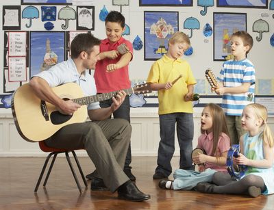 Links auf dem Bild ist ein junger Lehrer, der Gitarre spielt abgebildet. Neben ihm stehen drei Jungs und sitzen zwei Mädchen, die circa 8 Jahre alt sind. Sie begleiten den Lehrer mit verschiedene Instrumente, z.B. Klanghölzern, Rasseln und Tamburin.