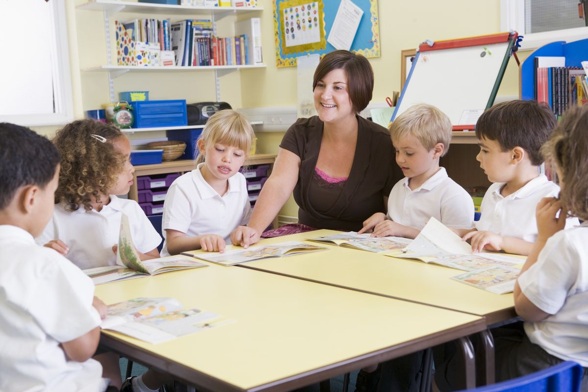 zur Vergrößerungsansicht des Bildes: Eine Grundschulleherin sitzt mit 6 Kindern an einem Tisch und bespricht einen Lesetext
