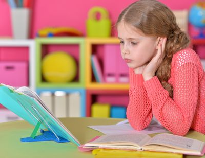 Auf dem Foto wird ein circa 9 Jahre altes Mädchen abgebildet, welches Hausaufgaben macht. Sie schaut in ein Buch und hat ihre Aufgaben vor sich liegen.