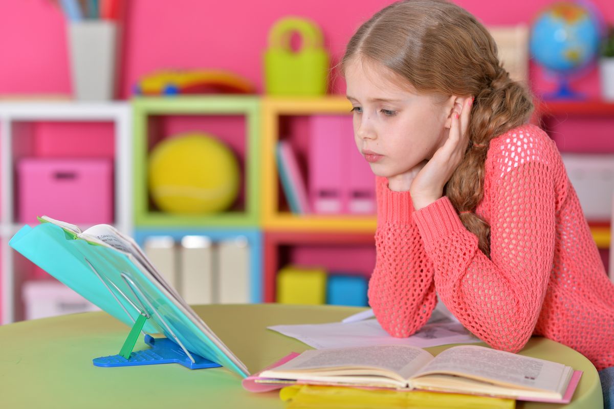 zur Vergrößerungsansicht des Bildes: Auf dem Foto wird ein circa 9 Jahre altes Mädchen abgebildet, welches Hausaufgaben macht. Sie schaut in ein Buch und hat ihre Aufgaben vor sich liegen.